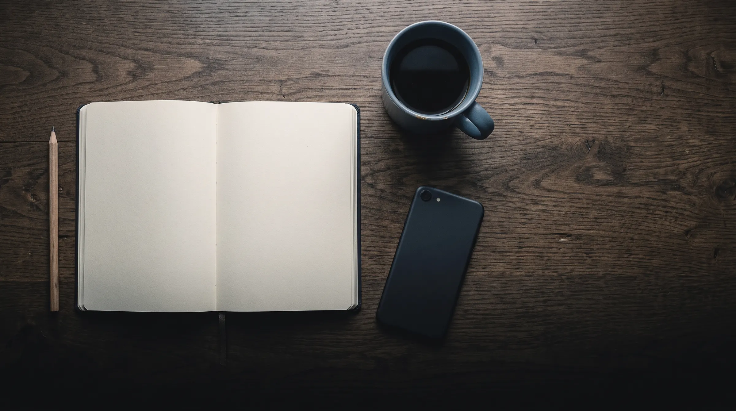 An open notebook with blank pages, a pencil, a cup of coffee and a phone on a wooden desk